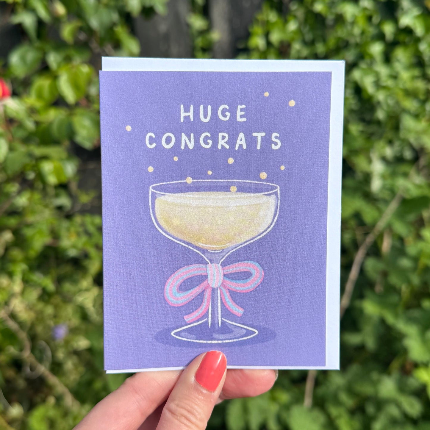 A hand with coral nail polish holds a lavender 'HUGE CONGRATS' greeting card featuring an illustration of a champagne coupe with a pink bow, against a blurred green garden background.